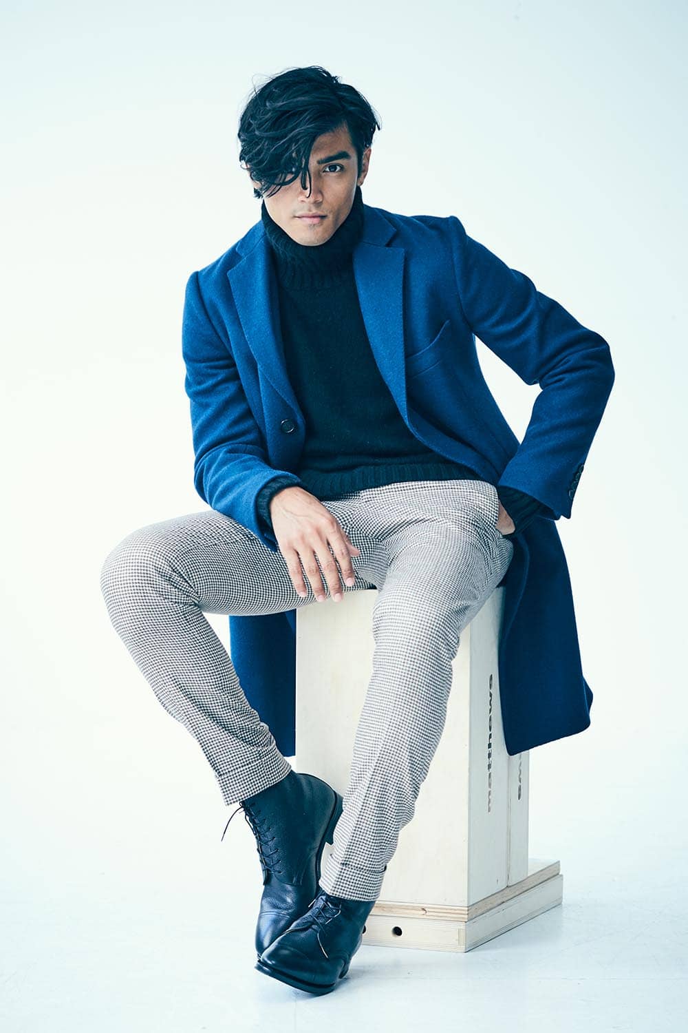 Ein Mann im blauem Mantel und karierter Hose sitzt auf einer Kiste